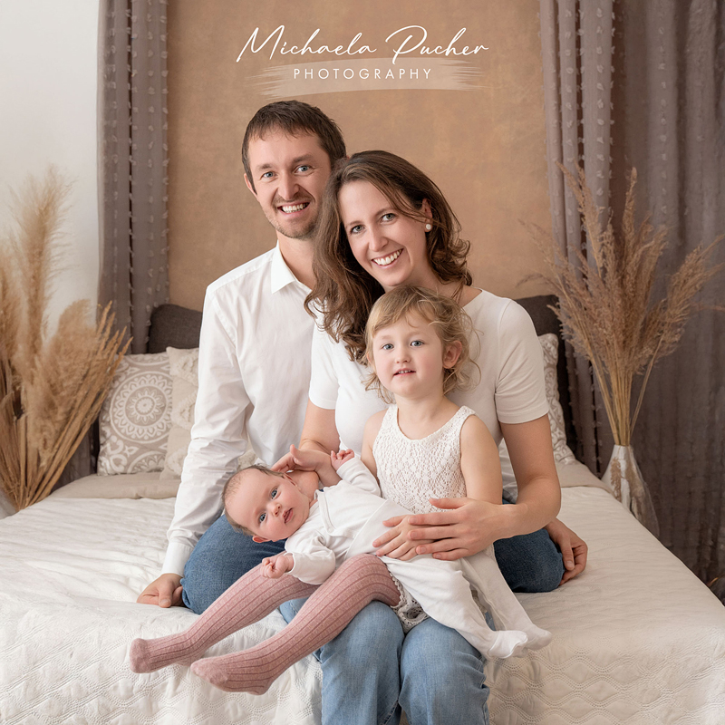 Eine vierköpfige Familie sitzt gemeinsam auf einem Bett in neutralen Naturtönen; das Baby liegt auf dem Schoß des Vaters und der großen Schwester, während alle in die Kamera lächeln.